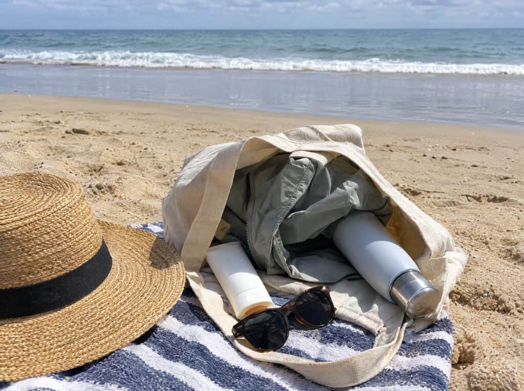 Beach tote with a straw hat, sunscreen, sunglasses and a rain jacket on the sand