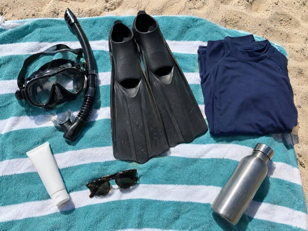 Snorkel mask, fins, rash guard, sunscreen and water bottle on a striped beach towel