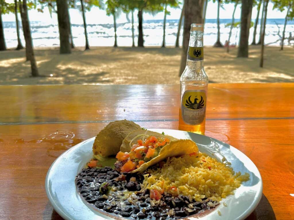 Plate of tacos with rice and beans and a beer at a beachfront table under palm trees